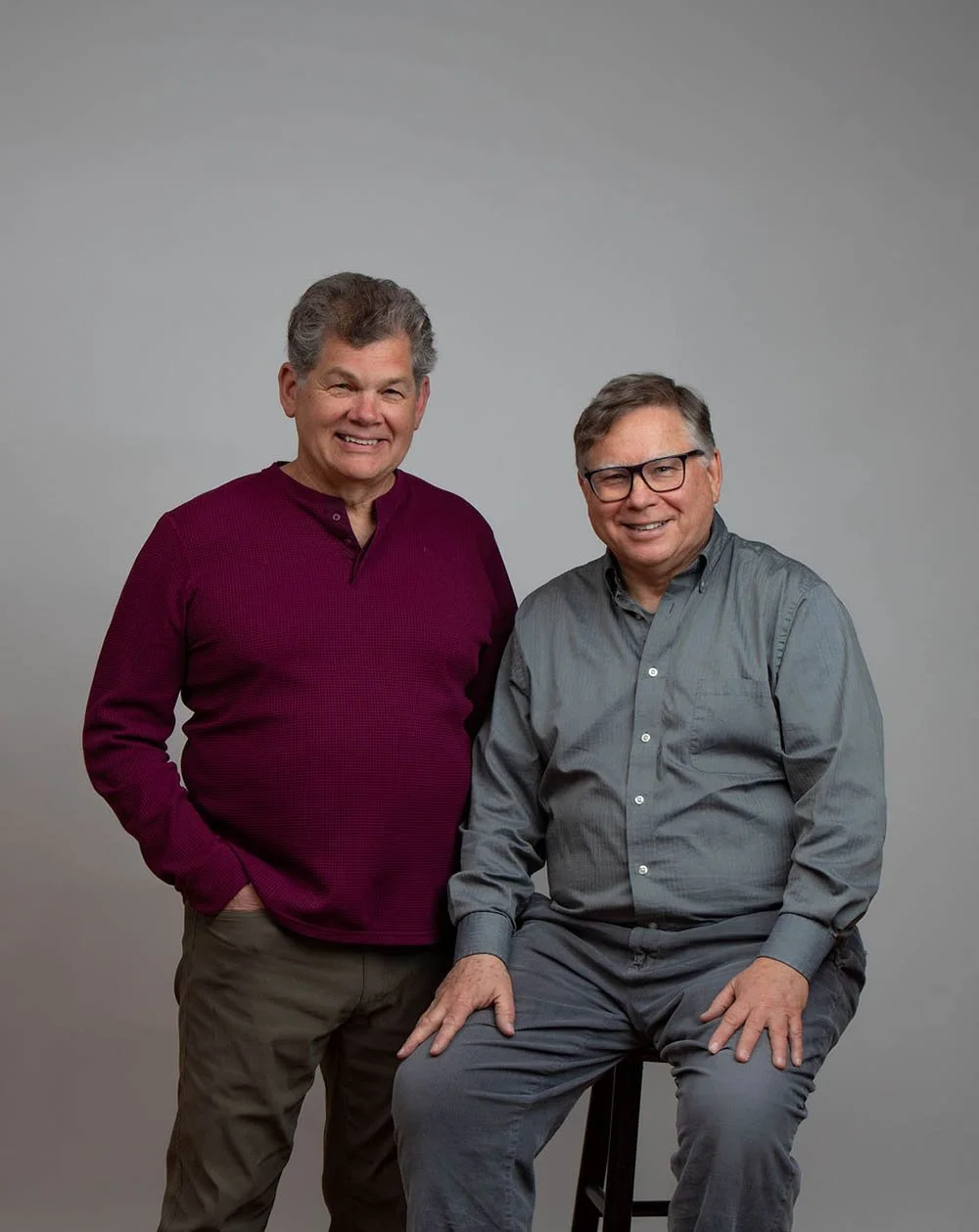 Two middle-aged men with glasses smiling pose against a plain gray background. One is standing with hands in pockets wearing a maroon sweater, and the other is sitting on a stool wearing a gray shirt and pants.