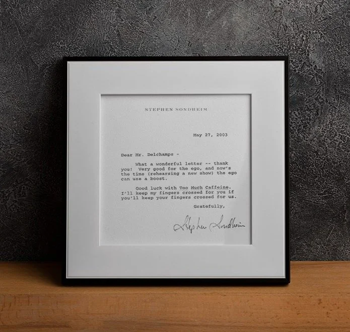 Framed letter on a wall, dated May 27, 2003, signed by Stephen Sondheim, with a dark textured wall background and a wooden surface beneath.