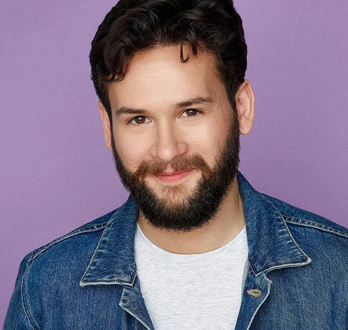 A young man with dark wavy hair and a full beard, smiling, wearing a denim jacket over a light gray t-shirt, against a purple background.