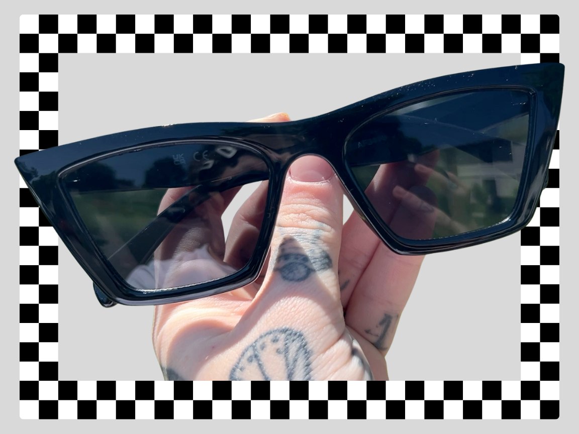 A person holding a pair of black sunglasses with rectangular lenses outdoors on a sunny day.