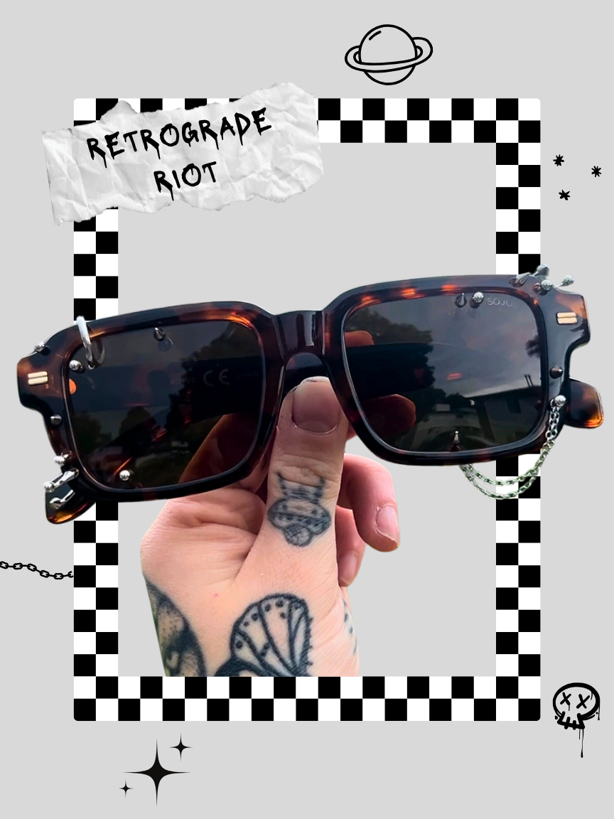 Person holding a pair of fashion sunglasses with tattoos on fingers, against a graphic background featuring a planet, stars, and a checkerboard border.