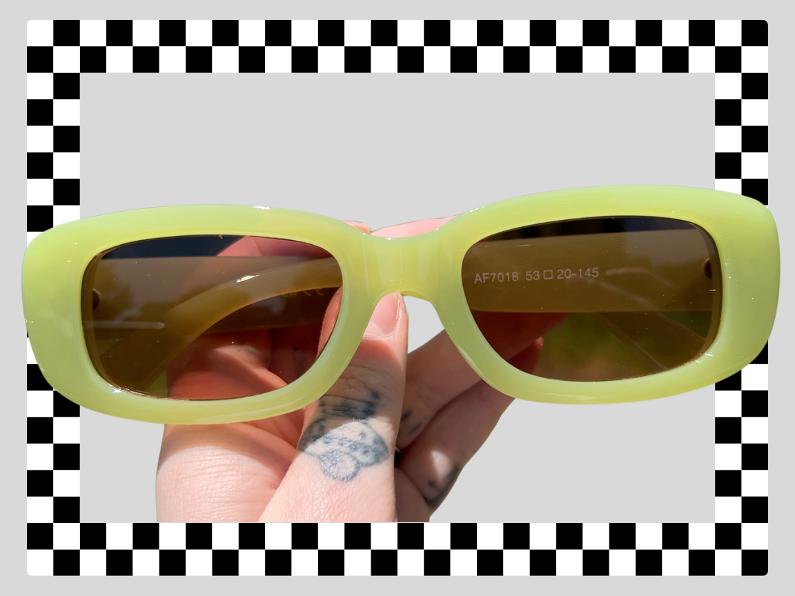 A pair of lime green sunglasses with dark lenses being held in a person's hand. The background is a checkered pattern with black and white squares, with a light gray rectangle in the center.