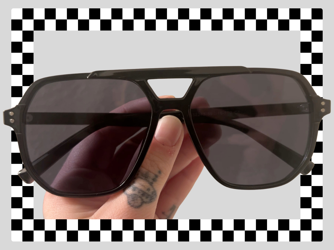 A hand holding black sunglasses with dark tinted lenses, set against a checkered background.