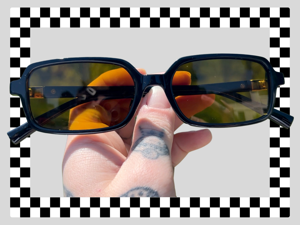 A hand with tattoos holding a pair of sunglasses with dark frames and tinted lenses, reflected with some outdoor scenery. The background includes a checkered black and white border.