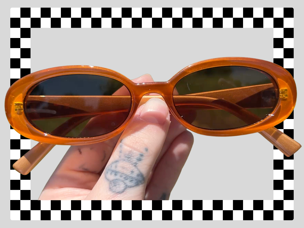 Person holding orange sunglasses with oval lenses and brown tinted glass, striped background with a checkerboard border.