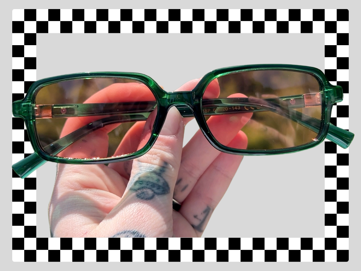 A hand with tattoos holding a pair of green sunglasses with tinted lenses against a gray background with a checkered border.