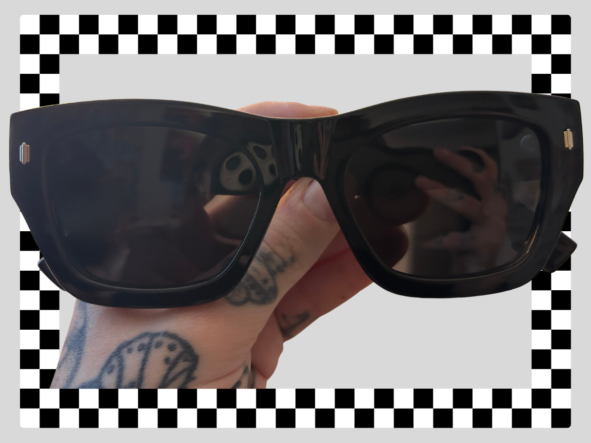 A person with tattoos holding a pair of black sunglasses with tinted lenses against a checkered background.