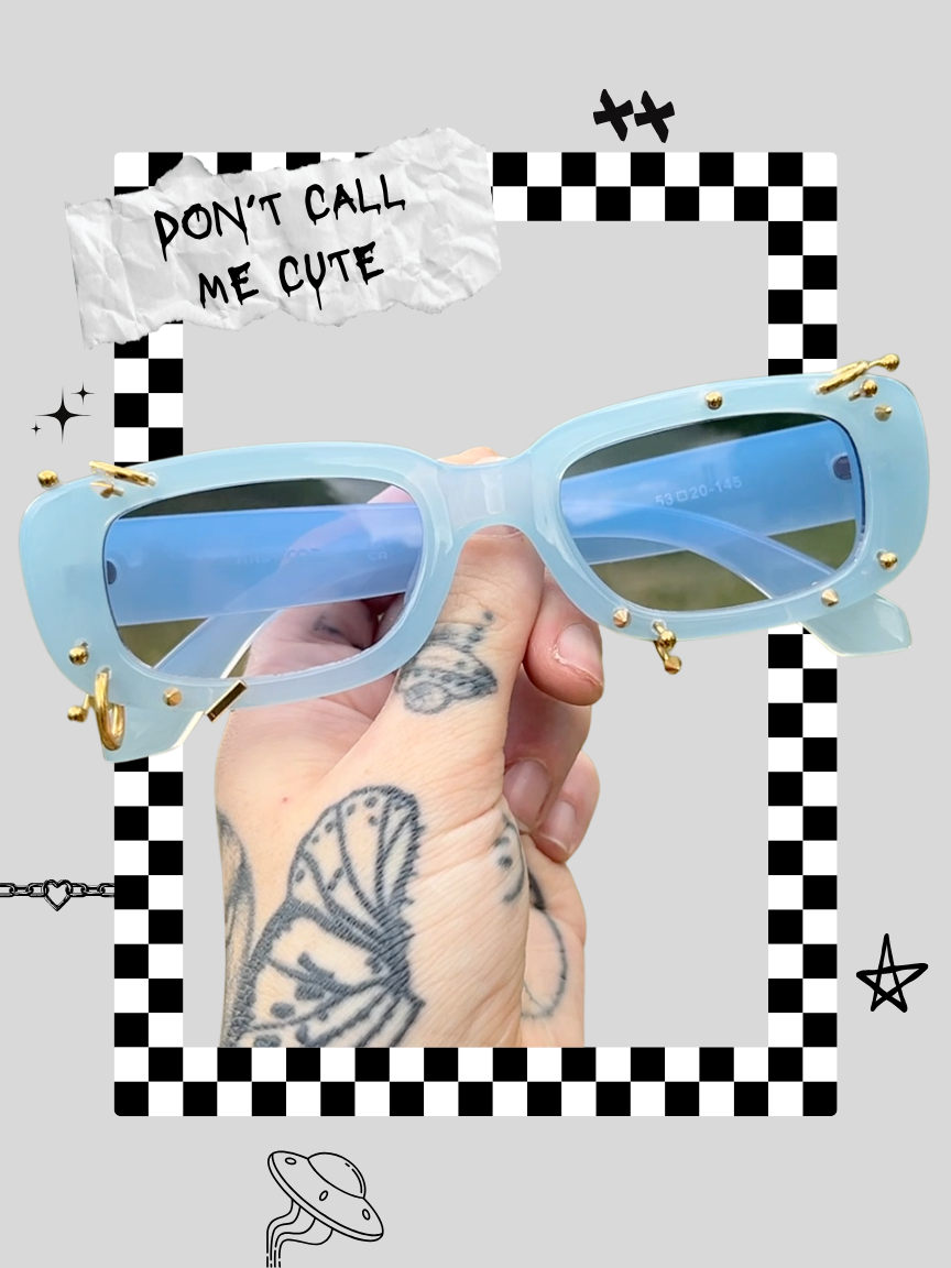 A person holding a pair of light blue sunglasses with gold accents in front of a decorative background featuring checkered patterns, doodles, and a torn paper sign that reads "DON'T CALL ME CUTE".
