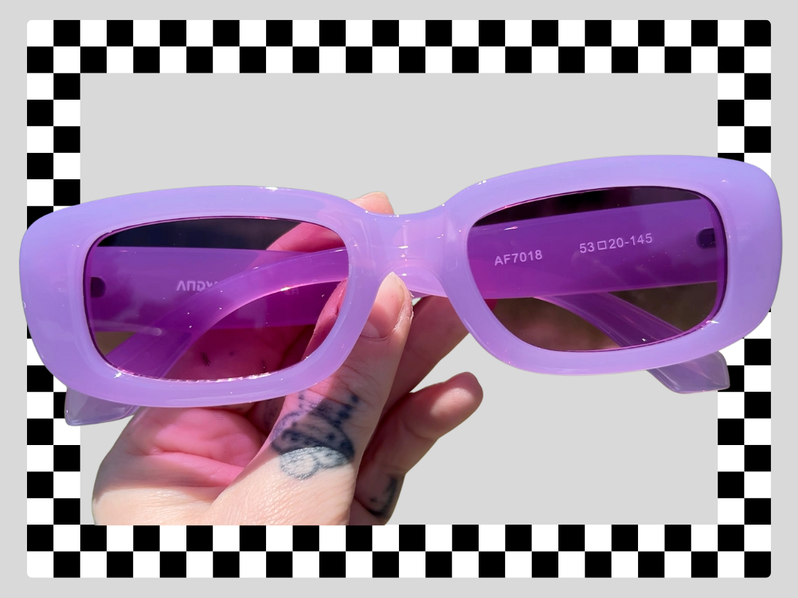 Purple sunglasses with tinted lenses being held by a hand with tattoos, against a background with a black and white checkered border.