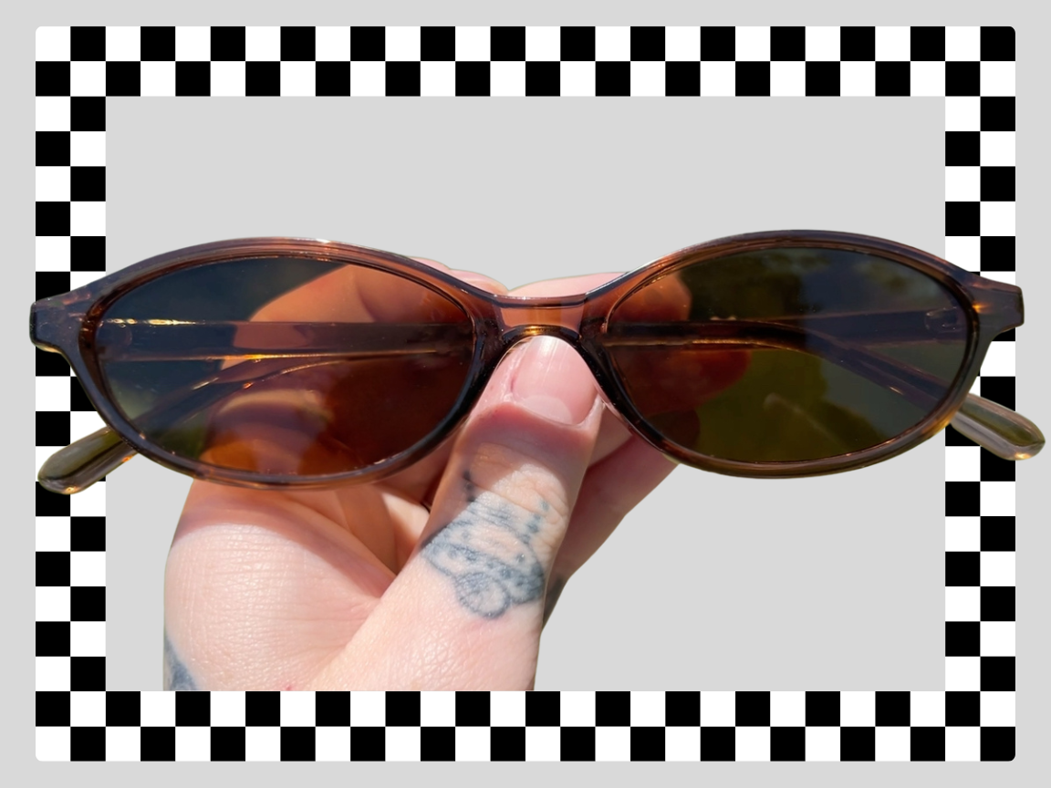 Person holding a pair of brown sunglasses with tinted lenses, tattooed hand, against a background with a checkered border.
