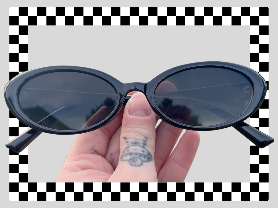 Close-up of a hand holding a pair of black oval sunglasses with dark lenses, with a checkerboard-patterned background.