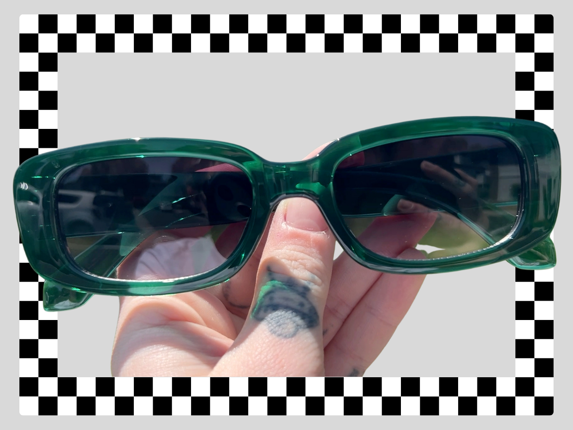Green rectangular sunglasses held by a person with tattoos on fingers; checkered border background.
