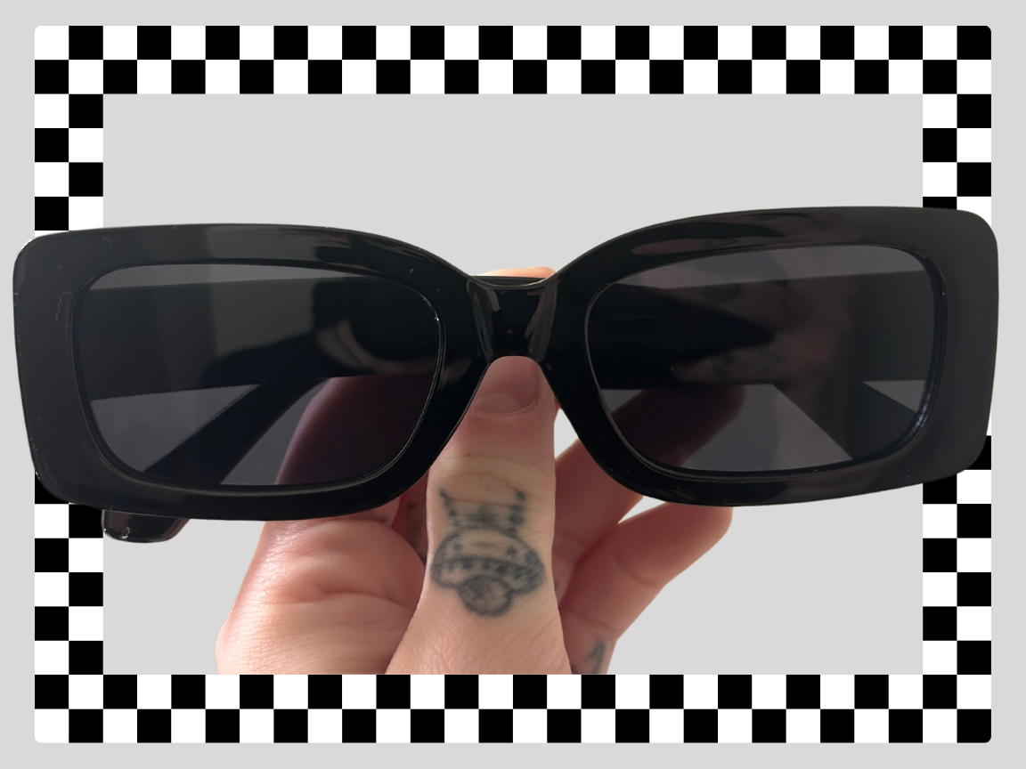 Person holding black sunglasses with dark lenses, tattooed finger, and checkered background.