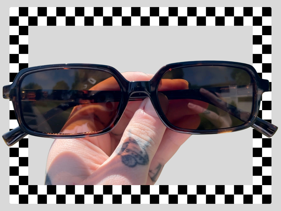 Hand holding black rectangular sunglasses with tinted lenses against an outdoor background.