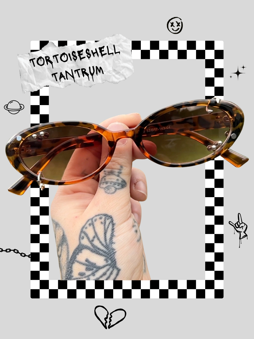 A hand with tattoos holding a pair of tortoise shell sunglasses with greenish lenses. The background features a checkered pattern and doodles of planets, a smiling face, a hand gesture, stars, and a broken heart, with a torn paper sign reading 'TORTOISESHELL TANTRUM'.
