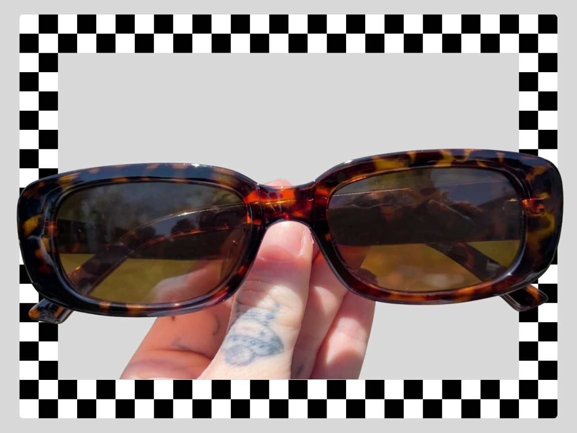 A person holding tortoiseshell sunglasses with dark lenses against a background of a checkered border and gray square.