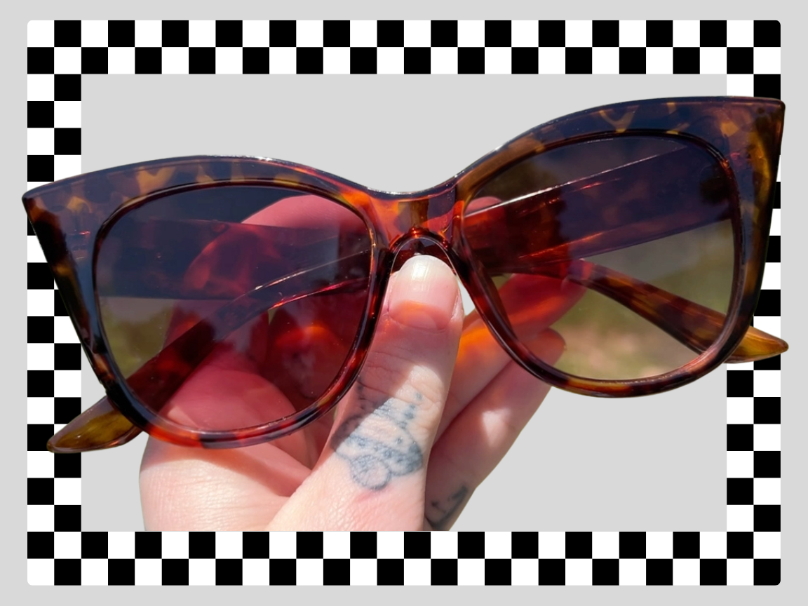 Close-up of a person's hand holding a pair of dark tortoise-shell sunglasses with a checkered border around the image.