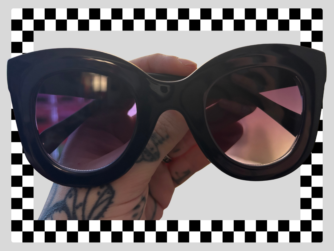 A person holding a pair of black sunglasses with pink-tinted lenses, tattoos visible on their hand, and a checkered black-and-white border around the image.