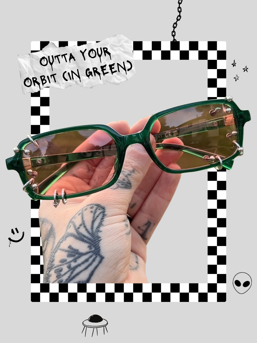 A person's hand with tattooed fingers holding a pair of green sunglasses with bronze accents. The background features a gray backdrop with a black and white checkered border, a torn note that reads "OUTTA YOUR ORBIT (IN GREEN)," a small alien face icon, a chain hanging from the top, and a doodle of a UFO at the bottom.