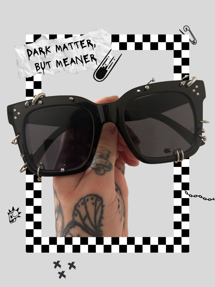 A person holding black sunglasses with metal spikes around the frame, surrounded by black and white checkered decorations, a written sign that says 'Dark Matter, But Meaner,' and various doodles like a skull, a chain, and shooting stars.
