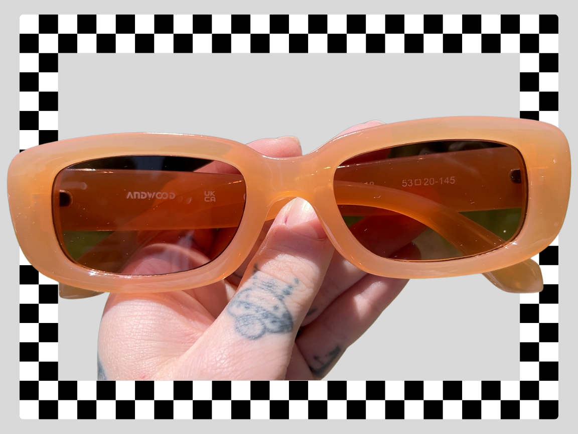 A hand holding orange sunglasses with dark lenses against a background with a black and white checkered border and gray interior.