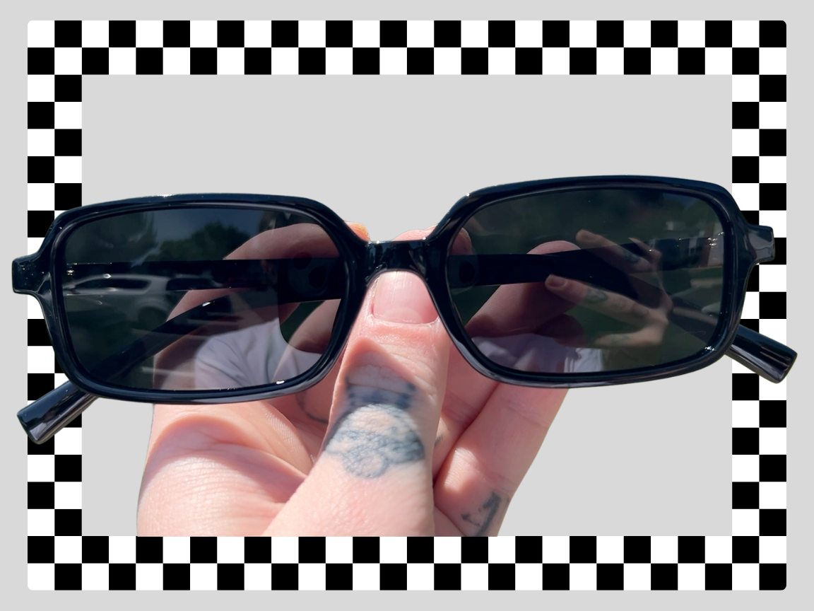 A hand holding black sunglasses with dark lenses outdoors, with a digital checkerboard border.
