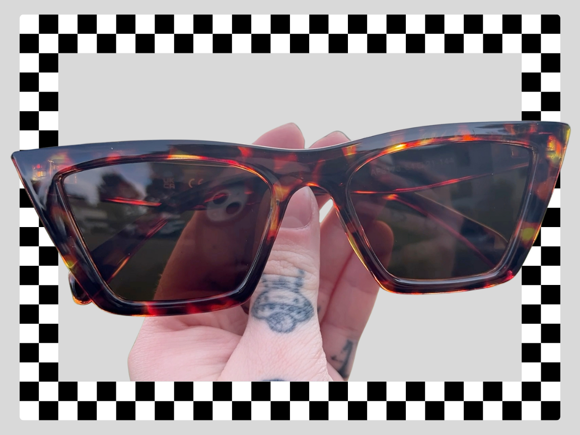Hand holding dark tortoiseshell sunglasses with black tinted lenses, black and white checkered border.