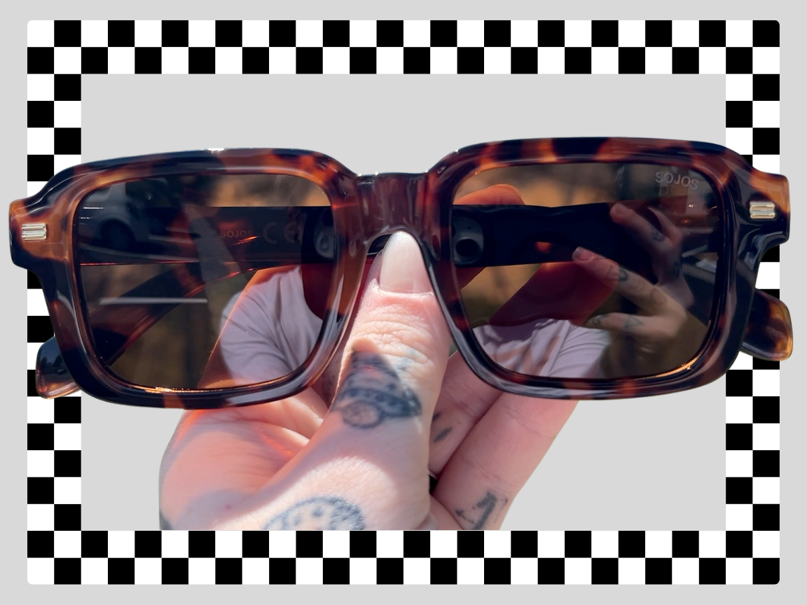 Person holding tortoise shell sunglasses with a hand tattooed with fingers and thumb visible, reflection of the person and background visible on the lenses, black and white checkered border and gray background.