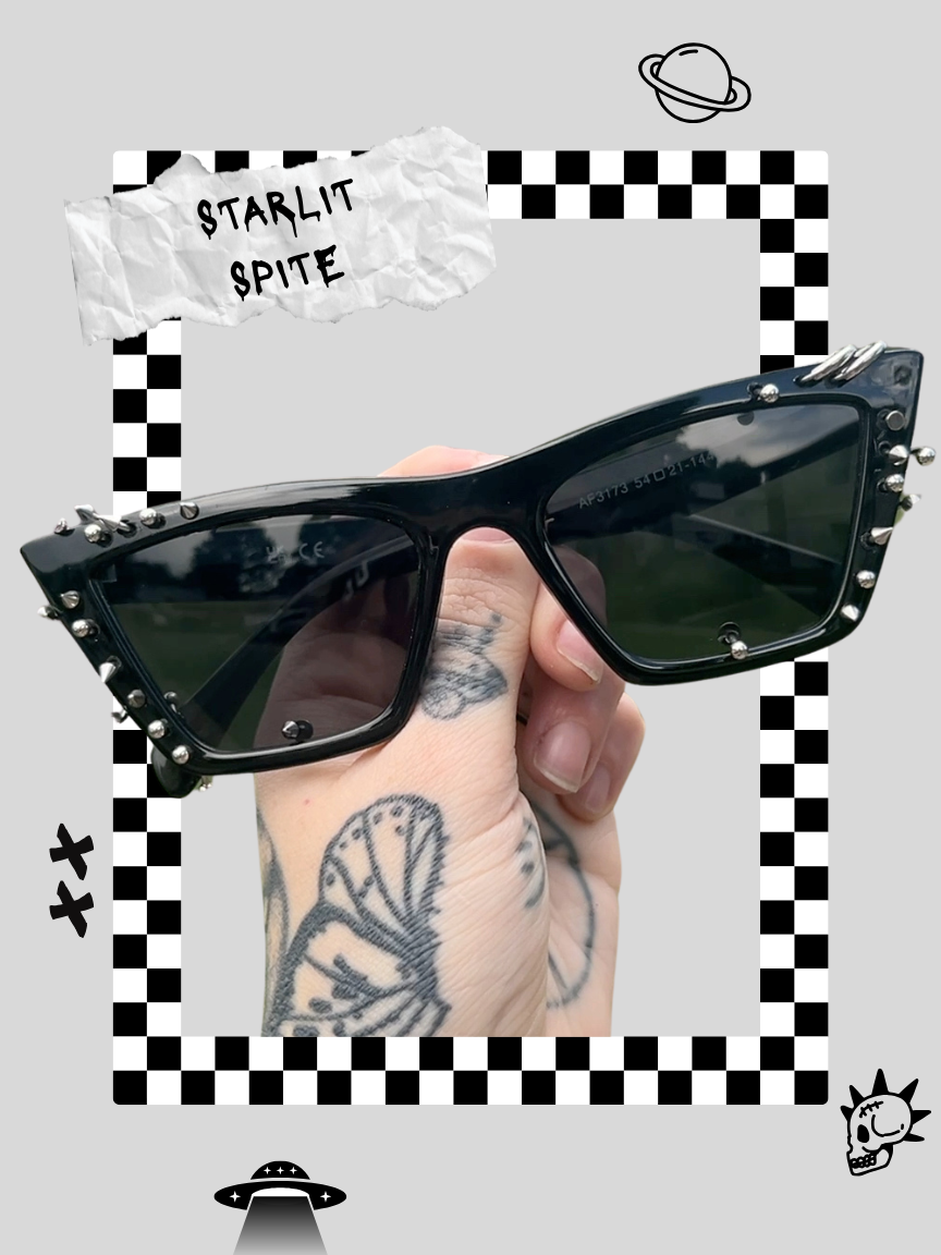 Hand holding black sunglasses with metal stud embellishments on the frame, against a gray background with doodles of a planet, a UFO, and a skull, and text "STARLIT SPITE" on a torn piece of paper.