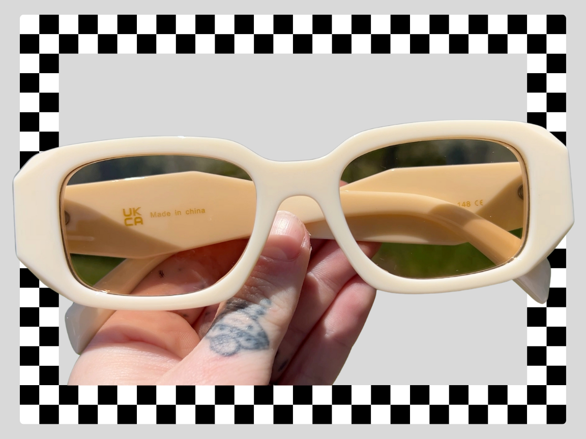 Beige sunglasses held by a hand with a tattooed finger, with a black and white checkered border