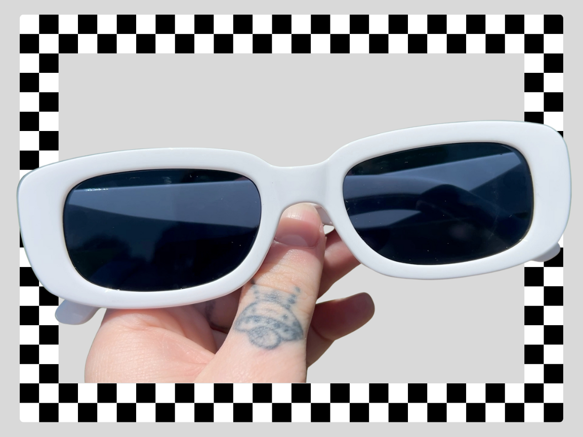 A person holding a pair of white sunglasses with dark lenses, with a tattoo finger, against a light background with a checkered border.