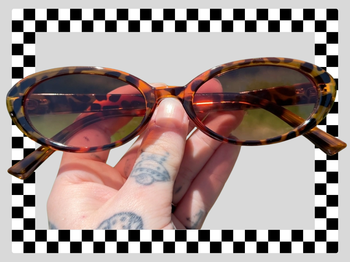 A person holding a pair of tortoise shell sunglasses with tinted lenses, with tattoos visible on their fingers, and an outdoor background reflected in the sunglasses.