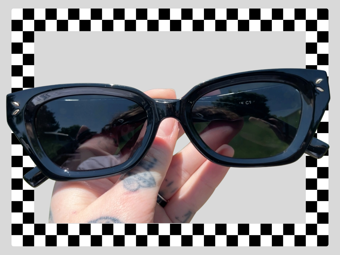 Black square-framed sunglasses with dark lenses held by a person with tattoos on their hand, reflection of a landscape in the lenses, and a checkered border around the image.