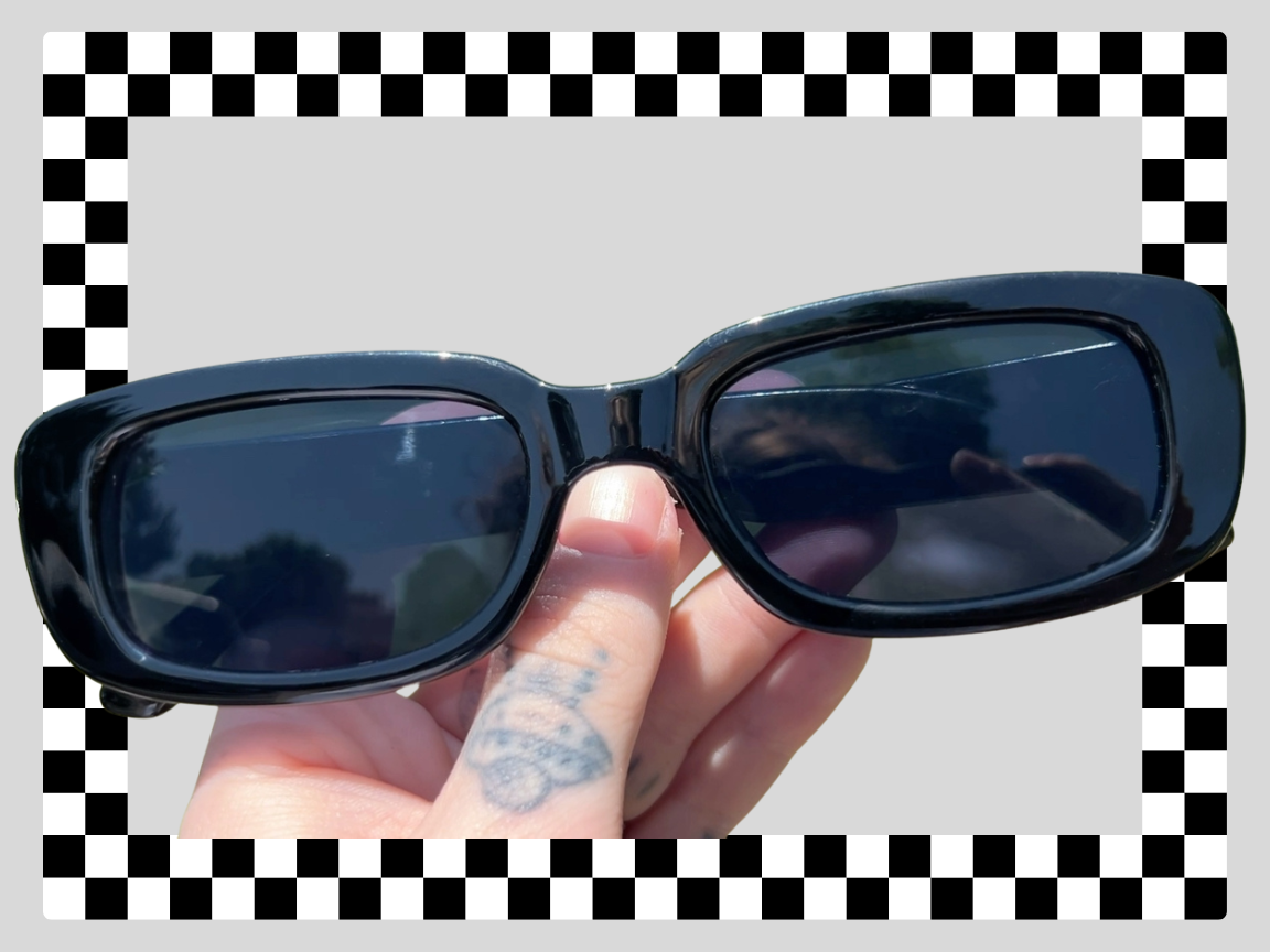 Close-up of a person holding black sunglasses with dark lenses outdoors, with a checkered background and a plain gray area behind the sunglasses.