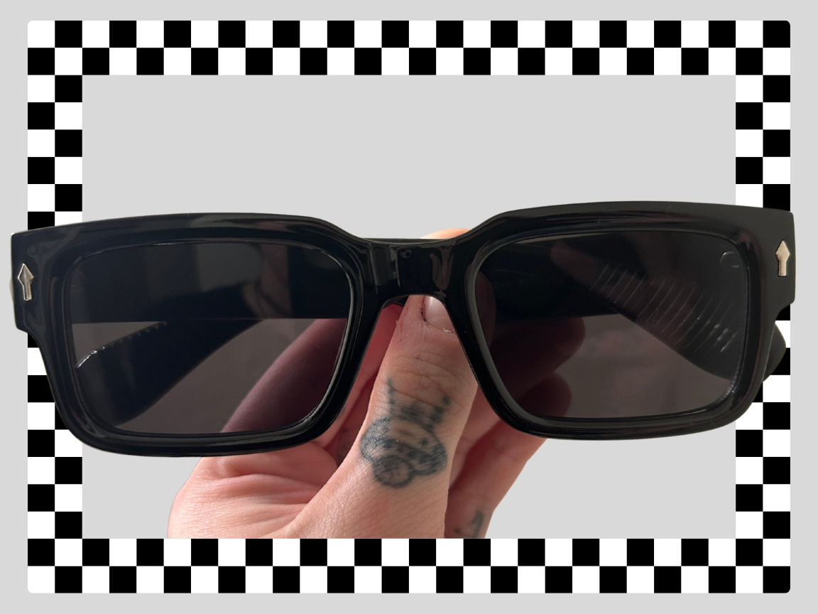 Black sunglasses with rectangular lenses held by a hand with a tattooed finger and knuckle. The background has a checkered and gray pattern.