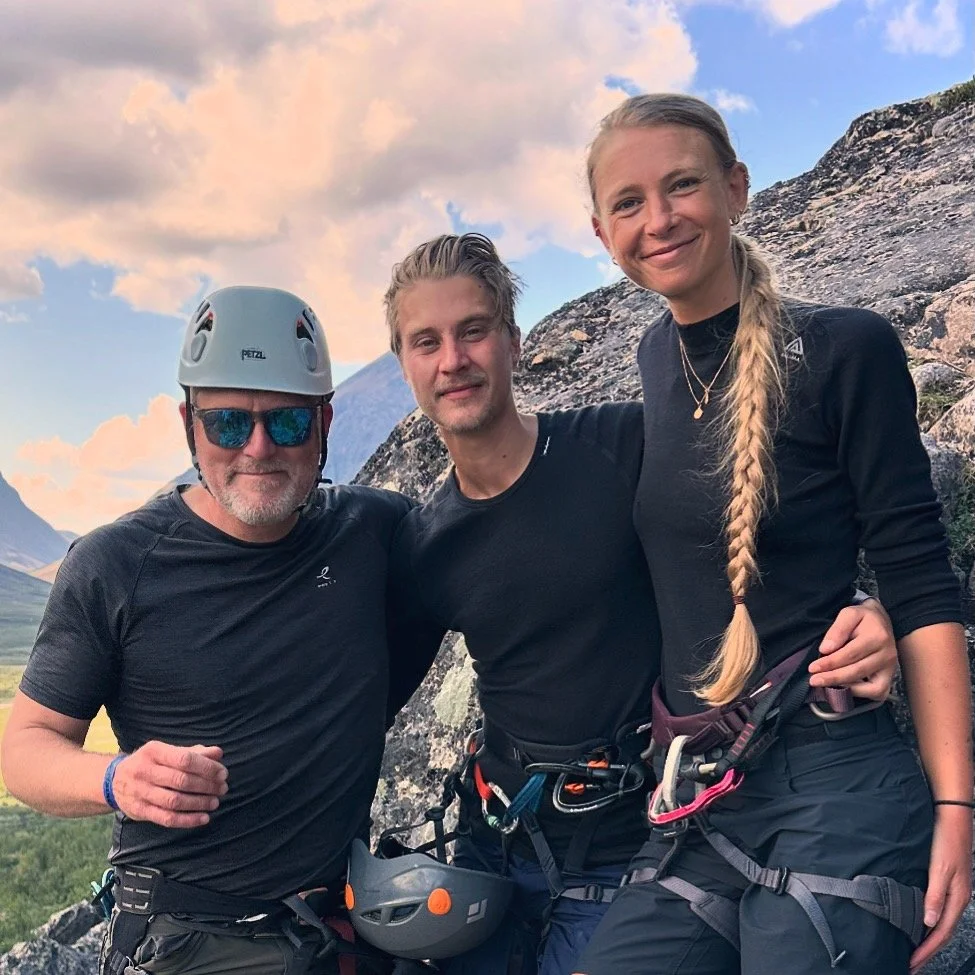 Tre personer som klättrar i bergsmiljö, två män och en kvinna, alla iklädda klättringsutrustning och kläder, med en bergskedja i bakgrunden.