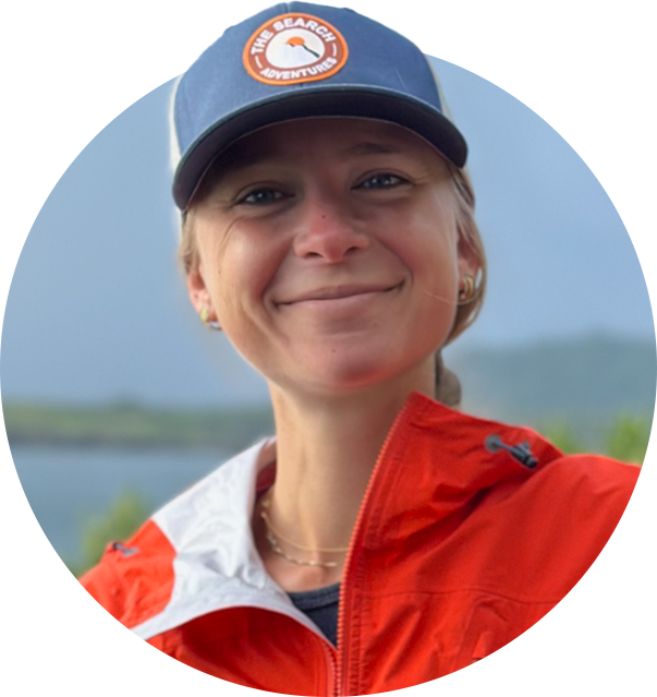 En kvinna med kort blont hår, som bär en blå keps med texten "The Search Adventure" och en röd jacka, utomhus med sjö och natur i bakgrunden.