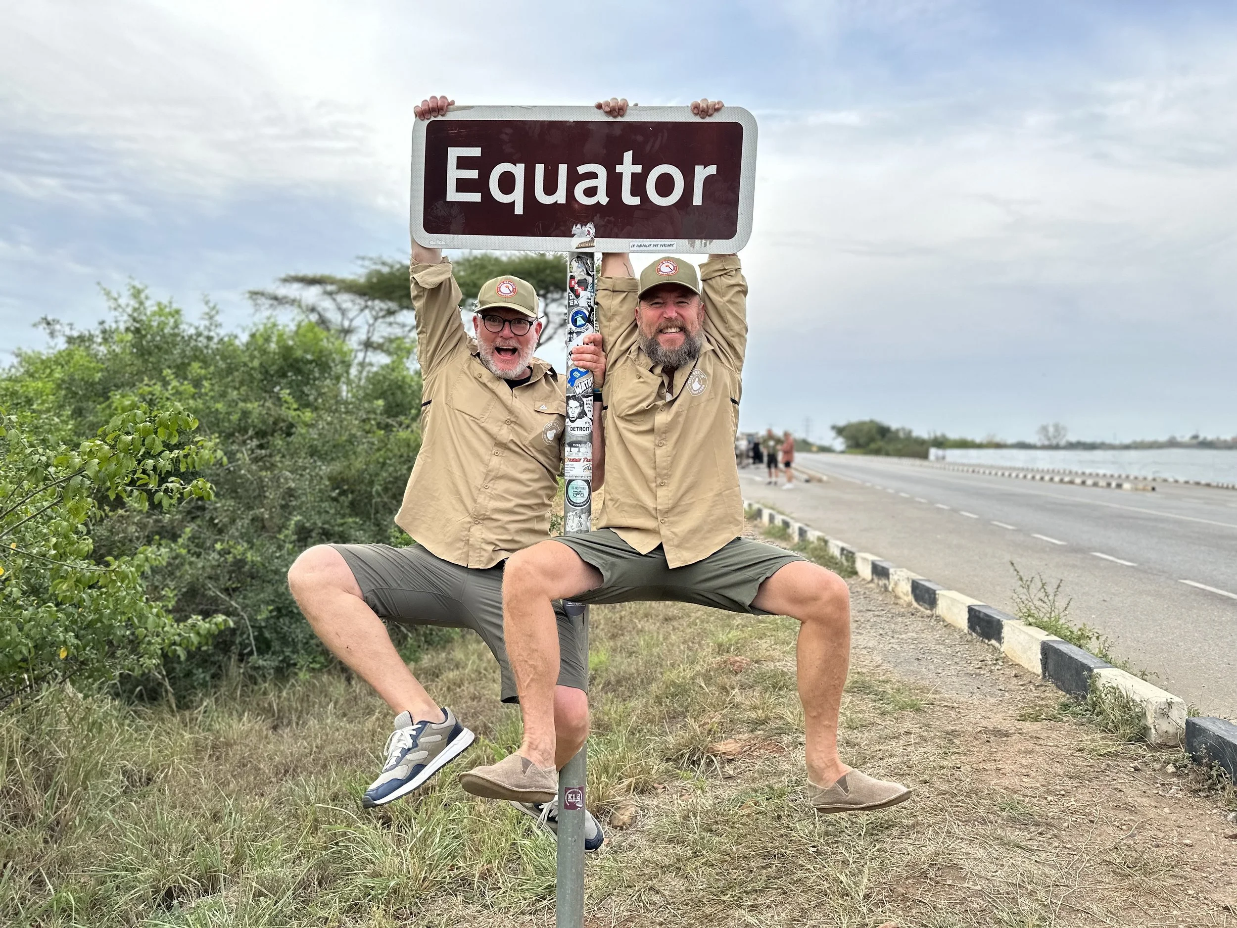 Två män i beige skjortor och kepsar ler och poserar framför ett vägmärke som säger 'Equator' vid ekvatorlinjen, med en väg och ett landskap i bakgrunden.