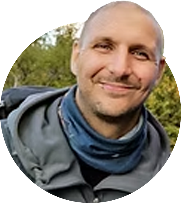 En man med kort hår och stubb ler, bär en grå jacka och en blå scarf, står utomhus med gröna träd i bakgrunden.