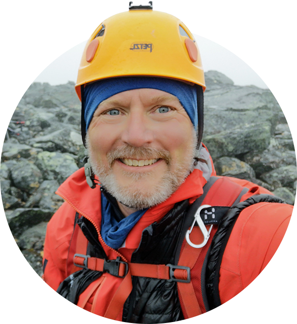 Man i klättrarutrustning tar en selfie på bergig terräng i grått landskap.