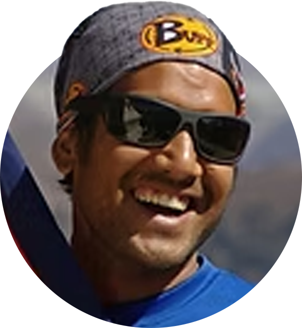 En glad man med solglasögon, svart och grått huvudband med en gul och svart logo, och en blå tröja, utomhus i bergsområde.