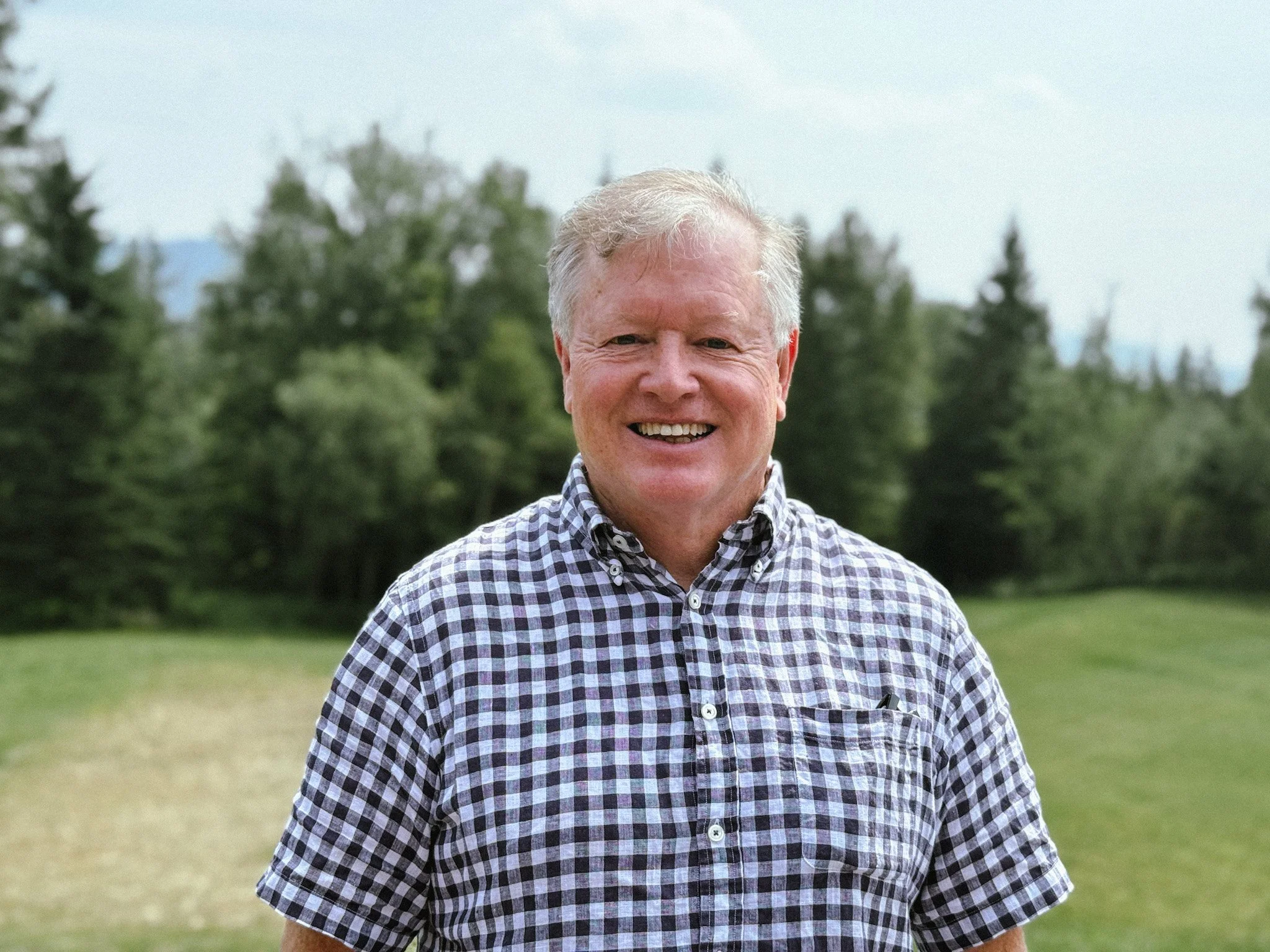 A smiling Craig McGowan with light hair wearing a checkered shirt standing outdoors in a grassy area with trees and cloudy sky in the background.