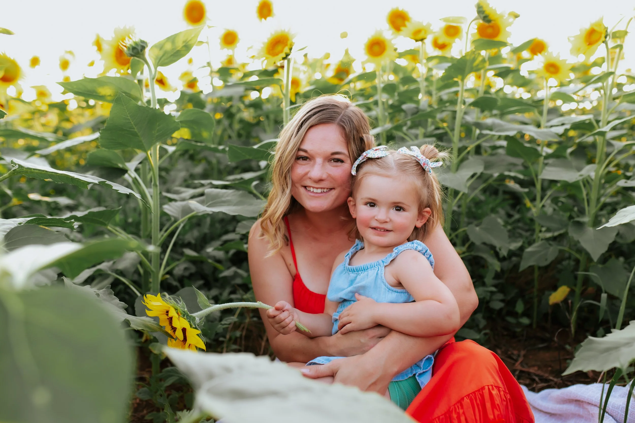 A woman and a young girl sitting in a sunflower field, smiling at the camera. The woman has wavy blonde hair and wears a red top; the girl has light brown hair tied with floral bows and wears a blue dress.