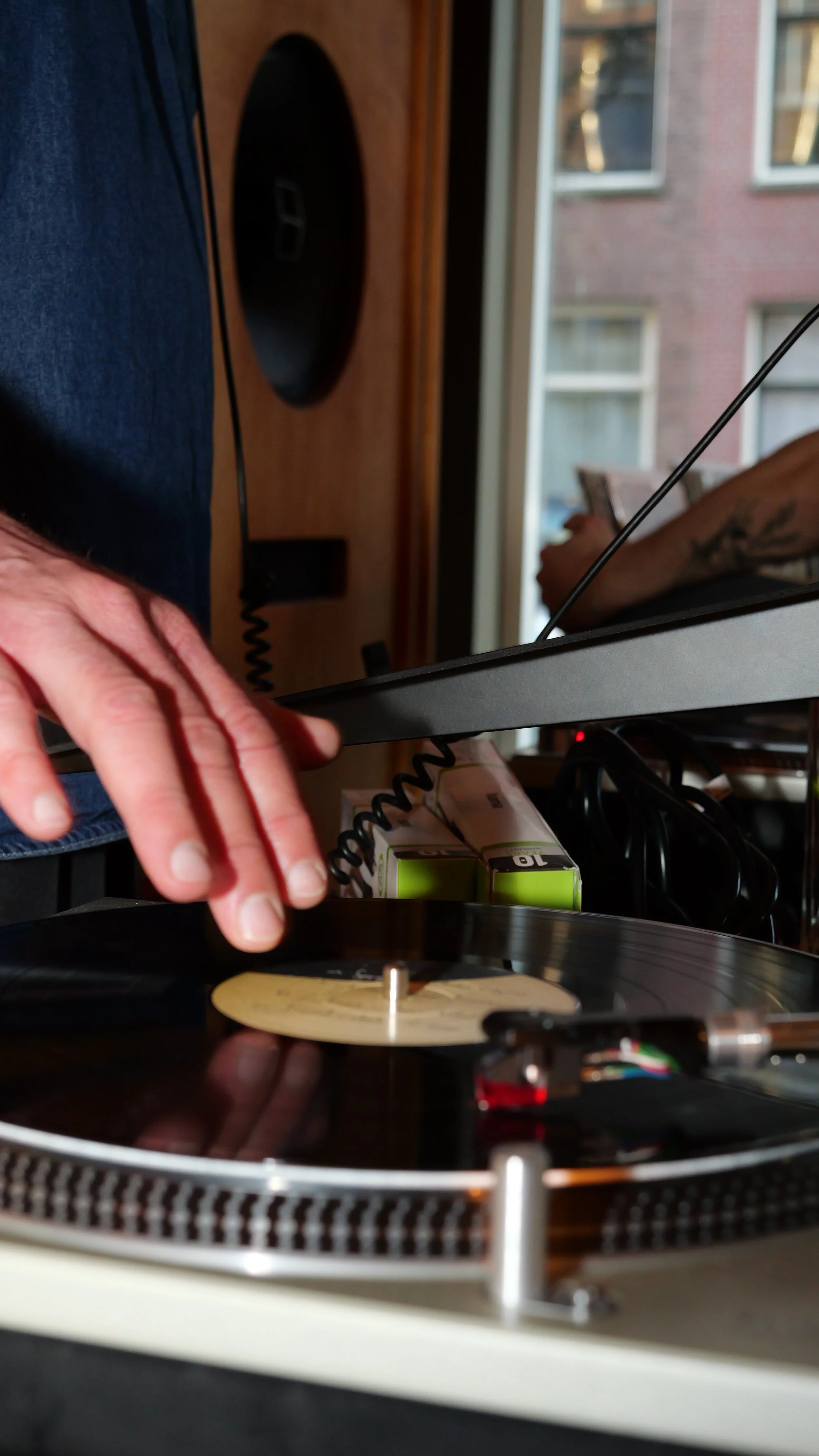 Person DJing with turntable and vinyl record, hand adjusting record on turntable, window in background.