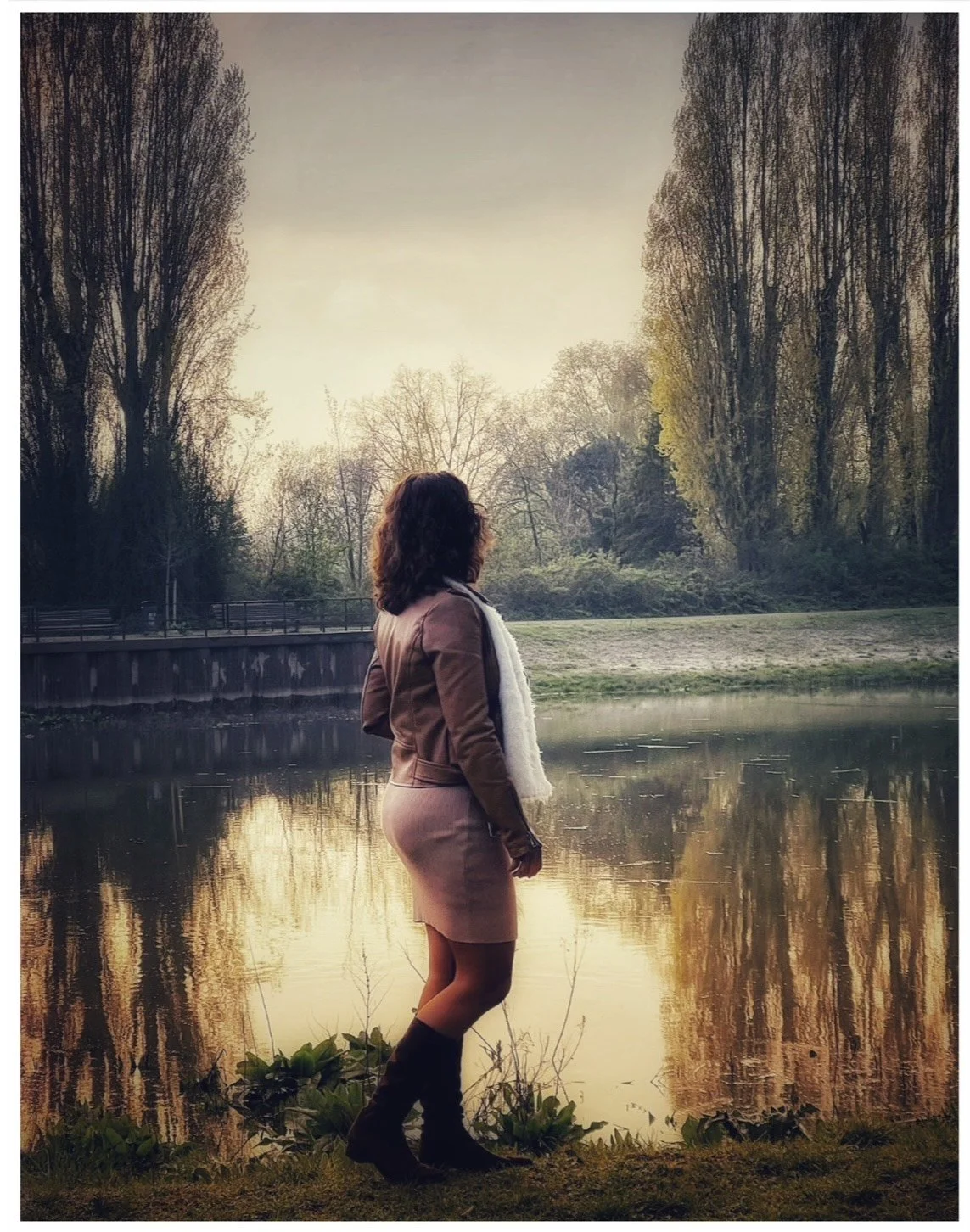 Woman in a brown jacket and skirt standing by a lake during sunset, with trees reflecting in the water.