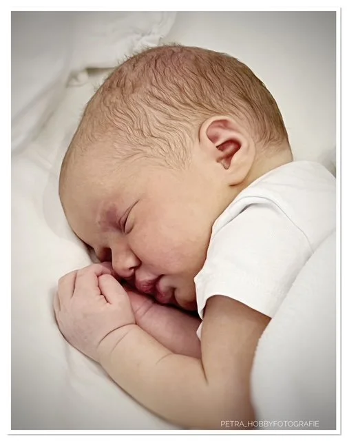 Close-up of a sleeping newborn baby with light brown hair, lying on a white pillow, using a white shirt, with hands near the face.