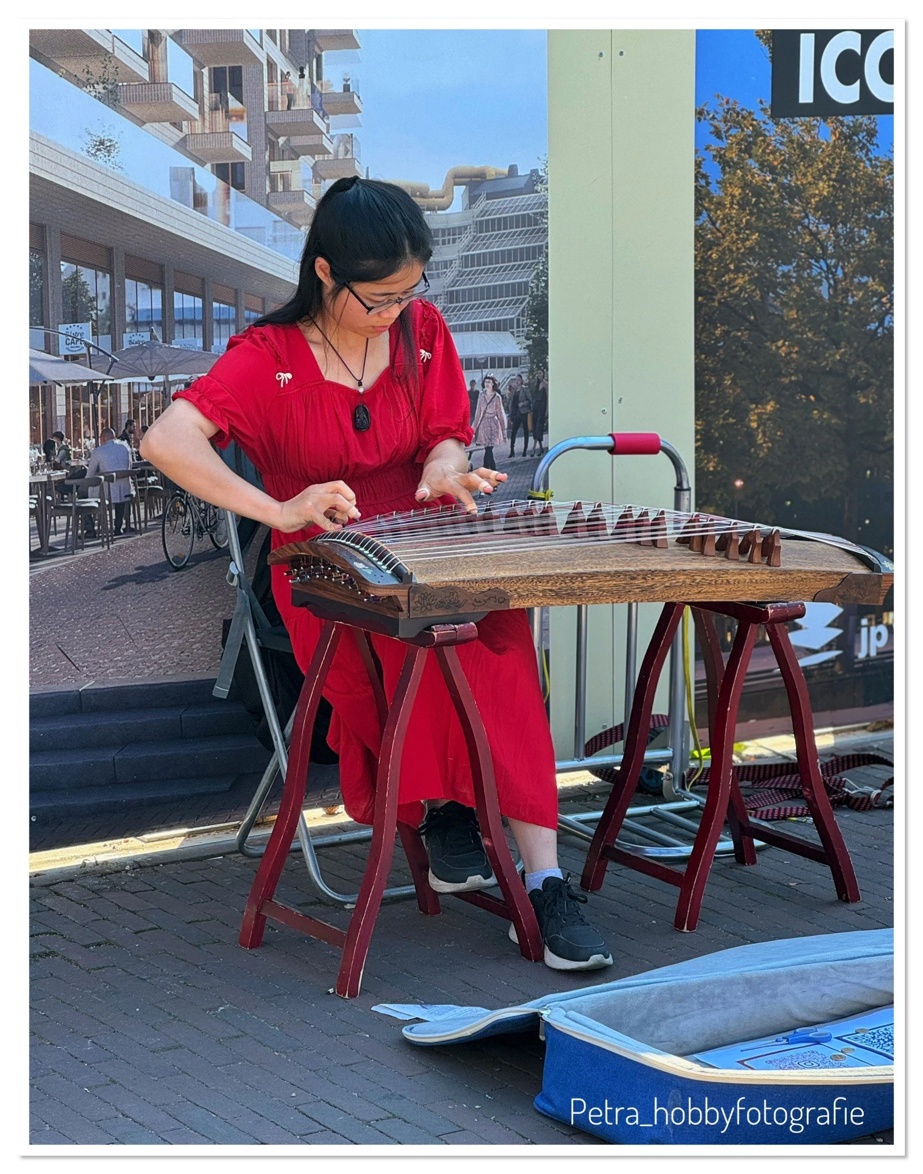 A woman in a red dress playing a wooden stringed instrument outdoors on a city sidewalk, with buildings, people, and outdoor cafes in the background.