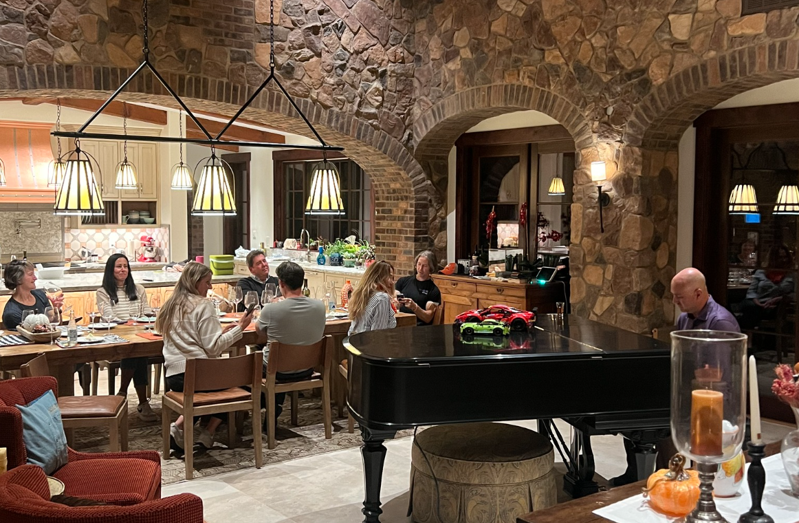 A group of people sitting around a dining table with wine glasses in a cozy, rustic kitchen with stone walls and arched brick doorways. There are hanging pendant lights over the table, and toy cars are on a black grand piano.