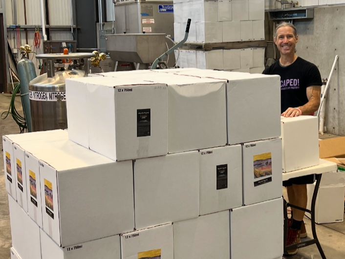 A man standing behind a stack of white boxes in a warehouse or industrial setting, with a smile on his face.