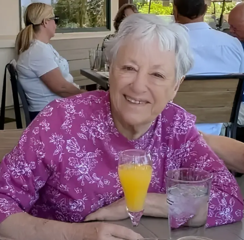 An elderly woman with white hair smiling at the camera, sitting at a table with a glass of orange juice and a glass of water, wearing a purple floral blouse, at an outdoor restaurant with other people in the background.